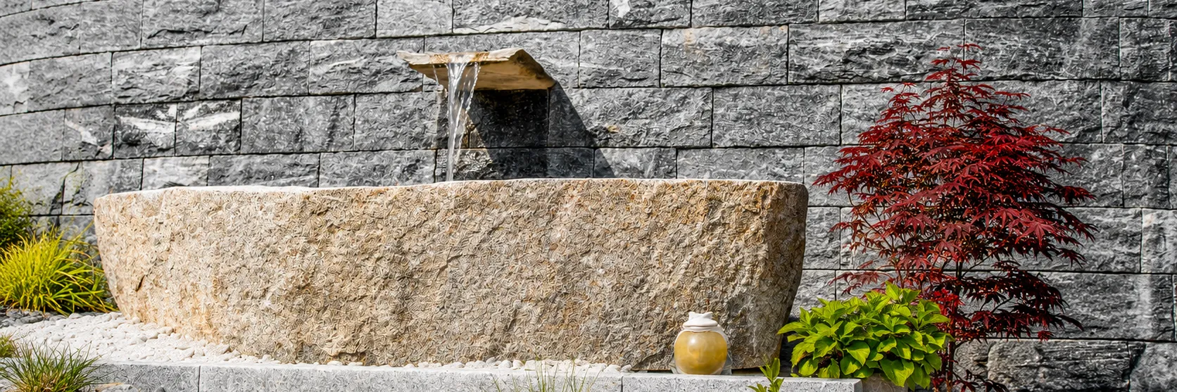 Wandbrunnen aus Naturstein mit Wasserauslauf, Granittrog und japanischem Ahorn vor heller Steinwand